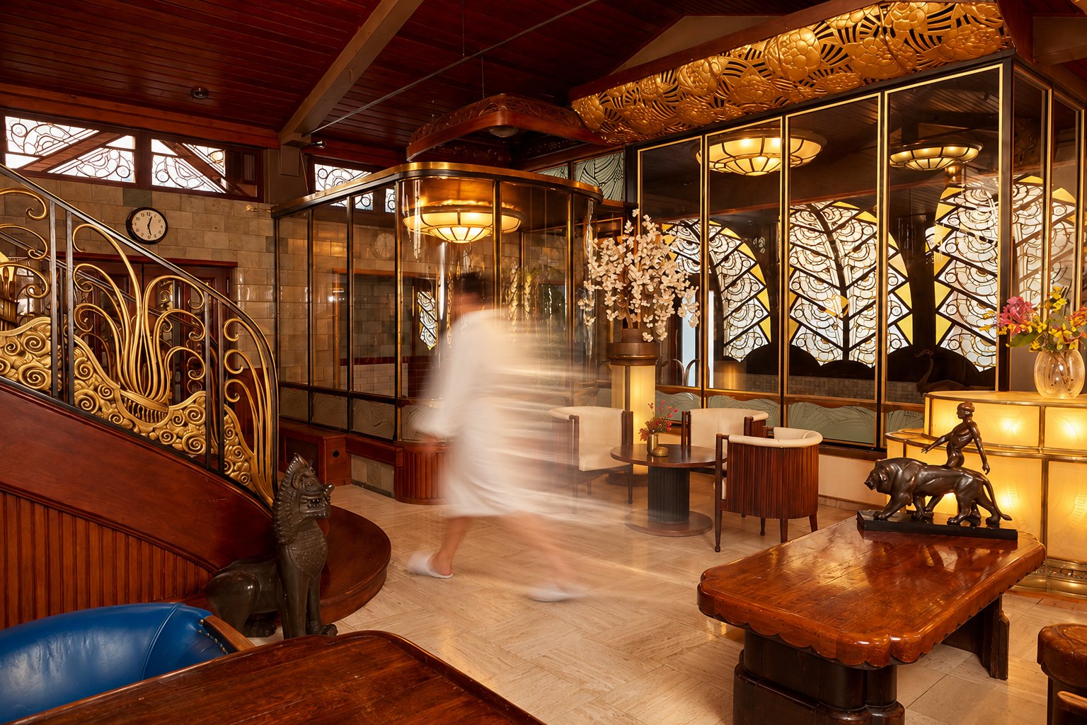 Een gast in badjas loopt door de lounge van Sauna Deco, langs verguld traphek, glas-in-lood en een houten leestafel met bronzen panter